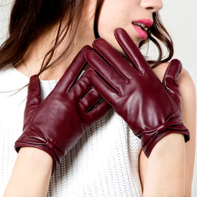 Photo de gants rouges en cuir avec petit noeud papillon portés par une femme brune au tee shirt blanc sans manche et tête tournée sur sa gauche, les mains à hauteur de son cou
