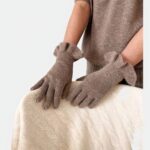 Photo d'une paire gants marron à volants en cachemire portés par une femme au pull de la même couleur à manches courtes, les mains gantées posées sur un pull en laine blanche torsadée, le tout sur un fond beige clair