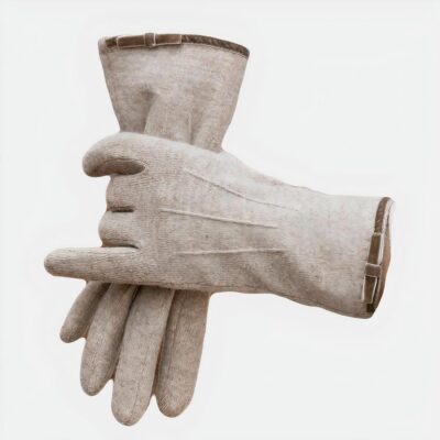 Photo représentant une main de femme dans un gant beige en laine cachemire à 3 surpiqures et ganse à noeud à l'ouverture, tenant l'autre gant entre ses doigts, la femme porte un manteau beige et se tient devant un meuble marron