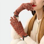 Une femme brune porte des gants orange avec des fleurs roses, et vertes. Une main qui touche sa bouche et l'autre touche l'autre poignet